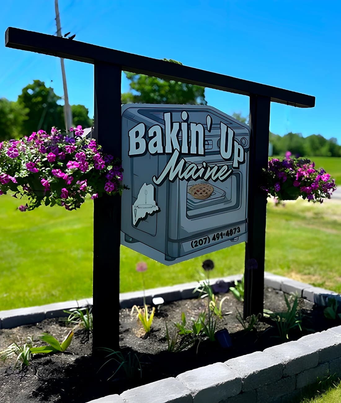 The Bakin' Up Maine roadside sign in Wilton, surrounded by flowers